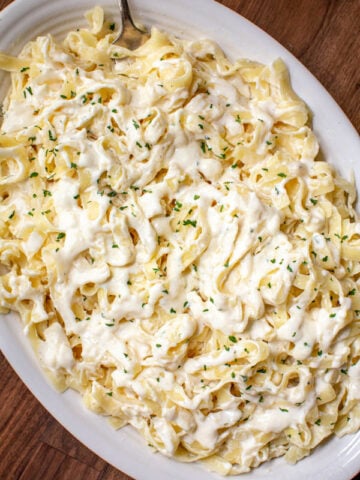 super creamy and rich fettuccine Alfredo sauce smothered in fettuccine pasta and parsley flakes on a white serving dish with a fork
