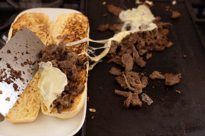image of Philly Cheesesteak filling being scooped inside a footlong bread roll with provolone cheese