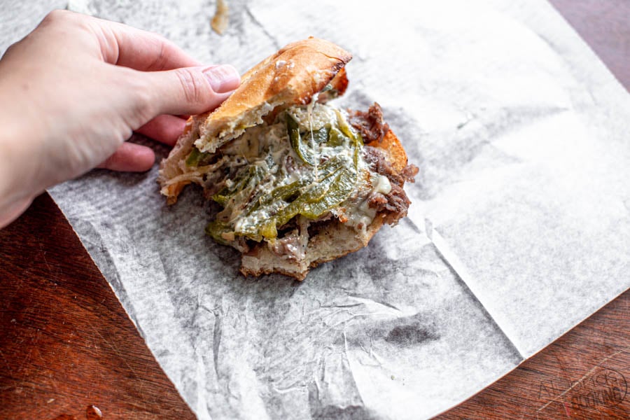 This Philly Cheesesteak Sandwich is made with marinated chuck roast shavings, caramelized onions, and peppers then broiled over toasted hoagie rolls with provolone cheese! It's one of the softest steak slices you'll have in a Philly Cheesesteak! Philly Cheesesteak Recipe How-to . #Phillycheesesteak #phillycheesesteakrecipe #cheesesteak #alyonascooking #phillycheesesteaksandwich #sandwich Philly Cheesesteak Sandwich on deli paper and bitten into