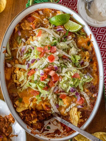 taco casserole in an oval baking pan with beef, beans, cheese, lettuce, tomato, and onion. Photographed with a lime wedge and sour cream and tortilla chips.