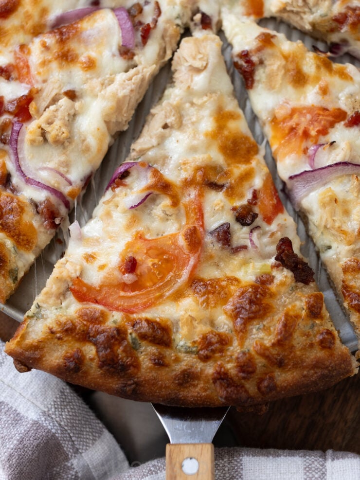 image of a whole wheat sourdough pizza slice zoomed in. Slice is topped with tomatoes, ranch dressing, mozzarella cheese, bacon bits, chicken, and red onion.