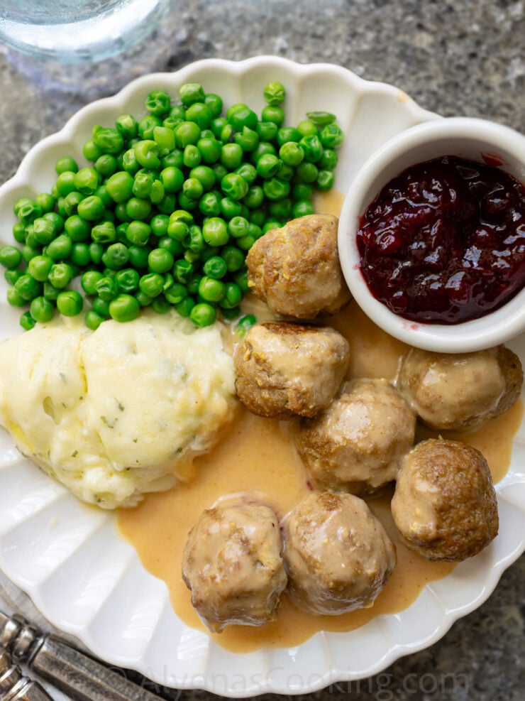 image of homemade Swedish meatballs smothered in a cream sauce and served with mashed potatoes, lingonberry sauce, and green peas (all plated on a scalloped white plate).
