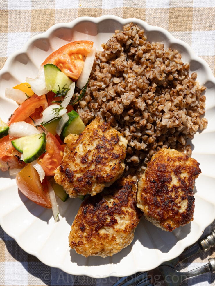 Ukrainian chicken Kotleti photographed outside by Alyona. It is plated with buckwheat and a fresh tomato and cucumber salad.