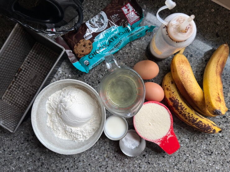 image of all the ingredients for chocolate chip banana bread (over-ripe bananas, vanilla extract, eggs, sugar, oil, milk, baking powder, baking soda, all-purpose flour, semi-sweet chocolate chips, and salt). Part of a 8x4 loaf pan and hand mixer is also photographed.