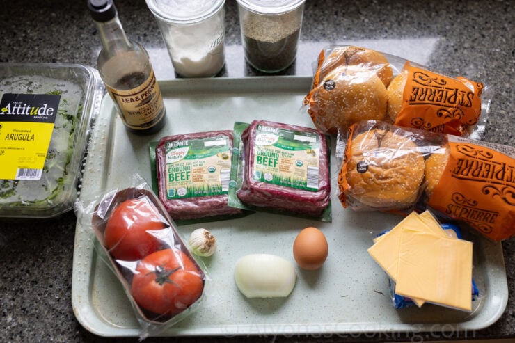 image of all the ingredients for oven baked burgers (hamburger buns, American cheese, onion, tomatoes, egg, garlic, ground beef, arugula, Worcestershire sauce, salt, and pepper).