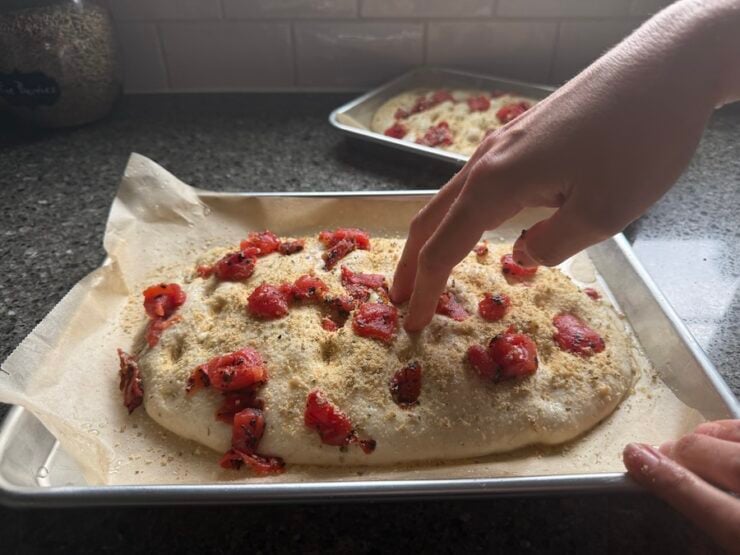 dimpling and making indents in the focaccia dough topped with parmesan herb topping and diced tomatoes