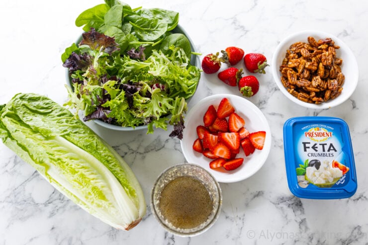 image of all the ingredients for spinach strawberry salad (one head of romaine lettuce, spring mix, baby spinach, fresh strawberries, homemade sweet and sour poppy seed dressing, feta cheese, and homemade maple candied pecans).